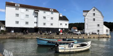 Two boats near Woodbridge Tidemill. The boats are fishing boats, one blue and one white. the mill is built of overlapping horizontal planks, painted white. There are some people sitting on benches on the quayside, and sme walking.