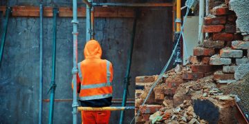 A construction worker in orange hi-viz stands with his back to the camera, leaning against some scaffolding on a building site