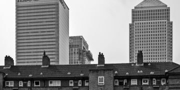 Gritty, black & white image capturing a striking contrast in London, juxtaposing traditional low-rise council flats in Tower Hamlets against the towering corporate skyscrapers of the Canary Wharf financial district.